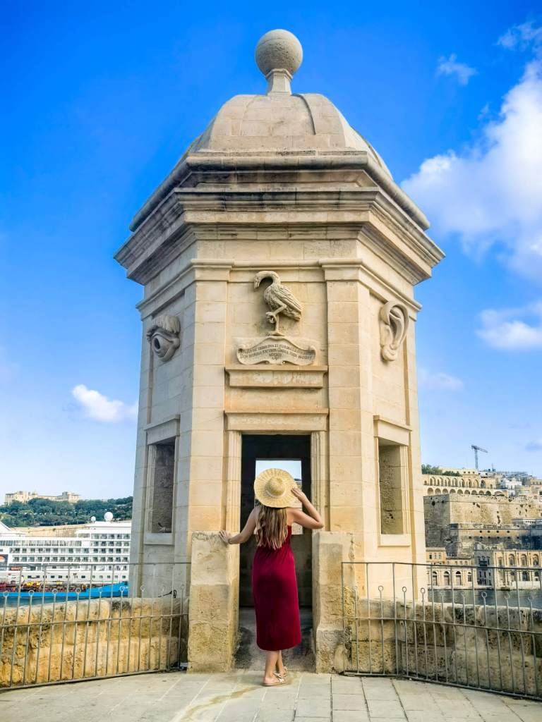 A Guide to Malta’s Three Cities: Discover Birgu, Isla and Bormla A woman in a red dress and straw hat looking out from the entrance of the Gardjola watchtower in Senglea, Malta.