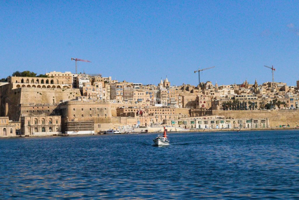 A Guide to Malta’s Three Cities: Discover Birgu, Isla and Bormla View of the Valletta city skyline across the blue water of the Grand Harbour with a small boat in the foreground.