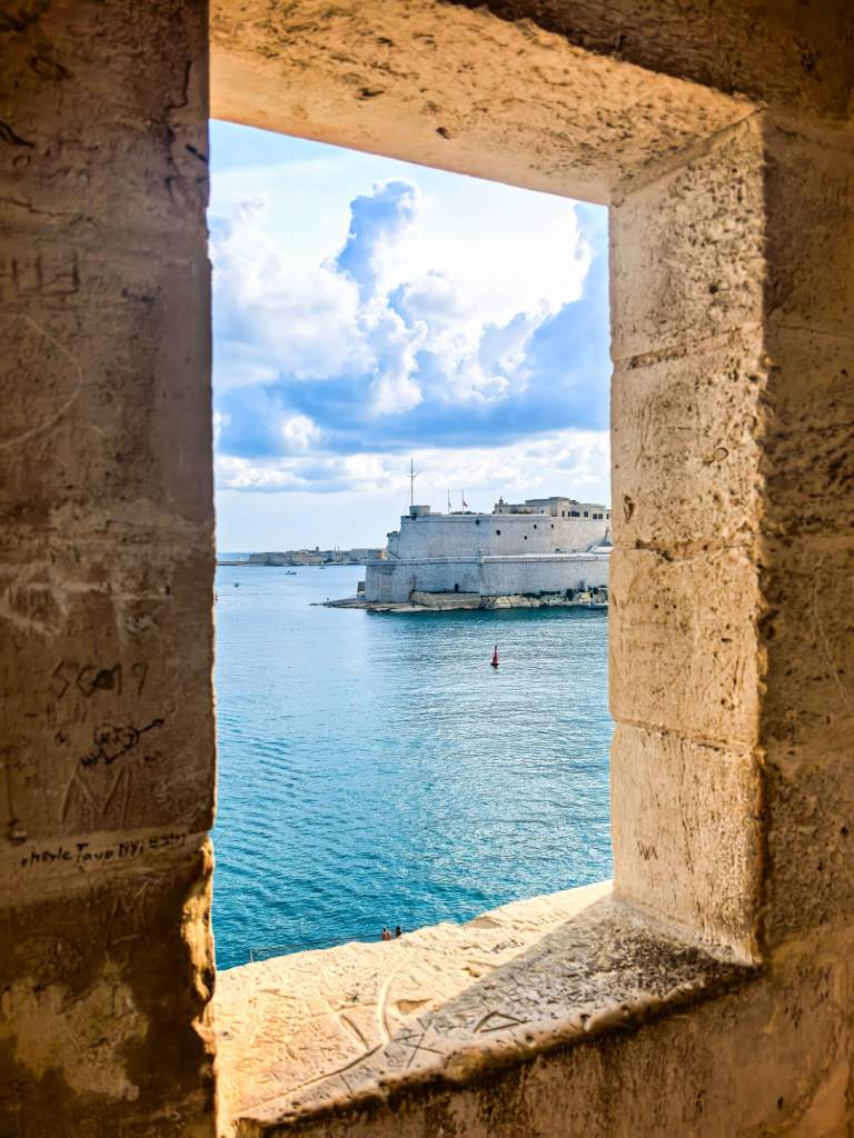 A Guide to Malta’s Three Cities: Discover Birgu, Isla and Bormla A view of Fort St Angelo across the blue water seen through a stone window in the bastions of the Three Cities.