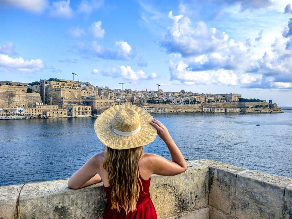 A Guide to Malta’s Three Cities: Discover Birgu, Isla and Bormla A woman in a red dress and straw hat looking at the Valletta skyline across the Grand Harbour from the Three Cities.