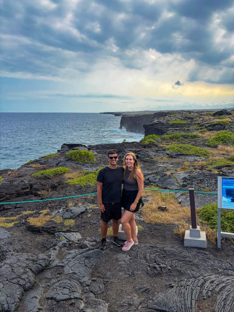 A couple standing on a rocky lava coastline by the ocean in Hawaii under a cloudy sky.