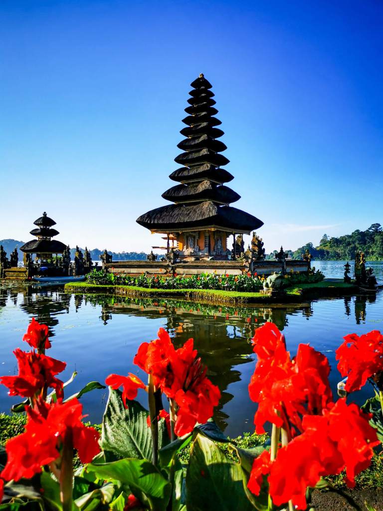 Pura Ulun Danu Bratan na Bali z czerwonymi kwiatami na pierwszym planie i odbiciem w wodzie.