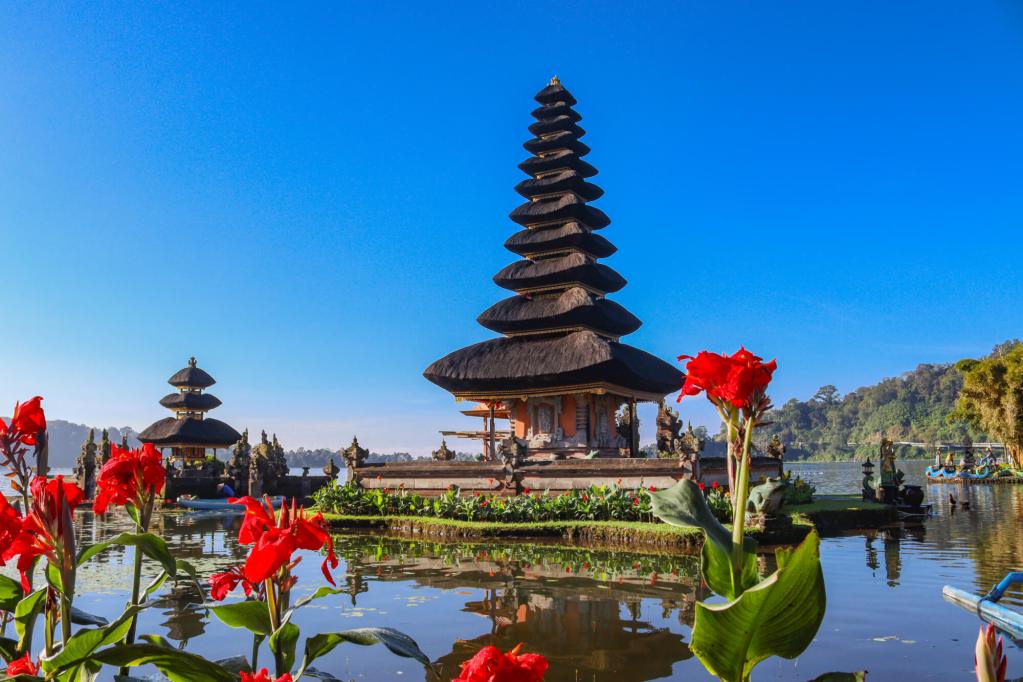 Pura Ulun Danu Bratan na Bali z czerwonymi kwiatami na pierwszym planie i odbiciem świątyni w wodzie.